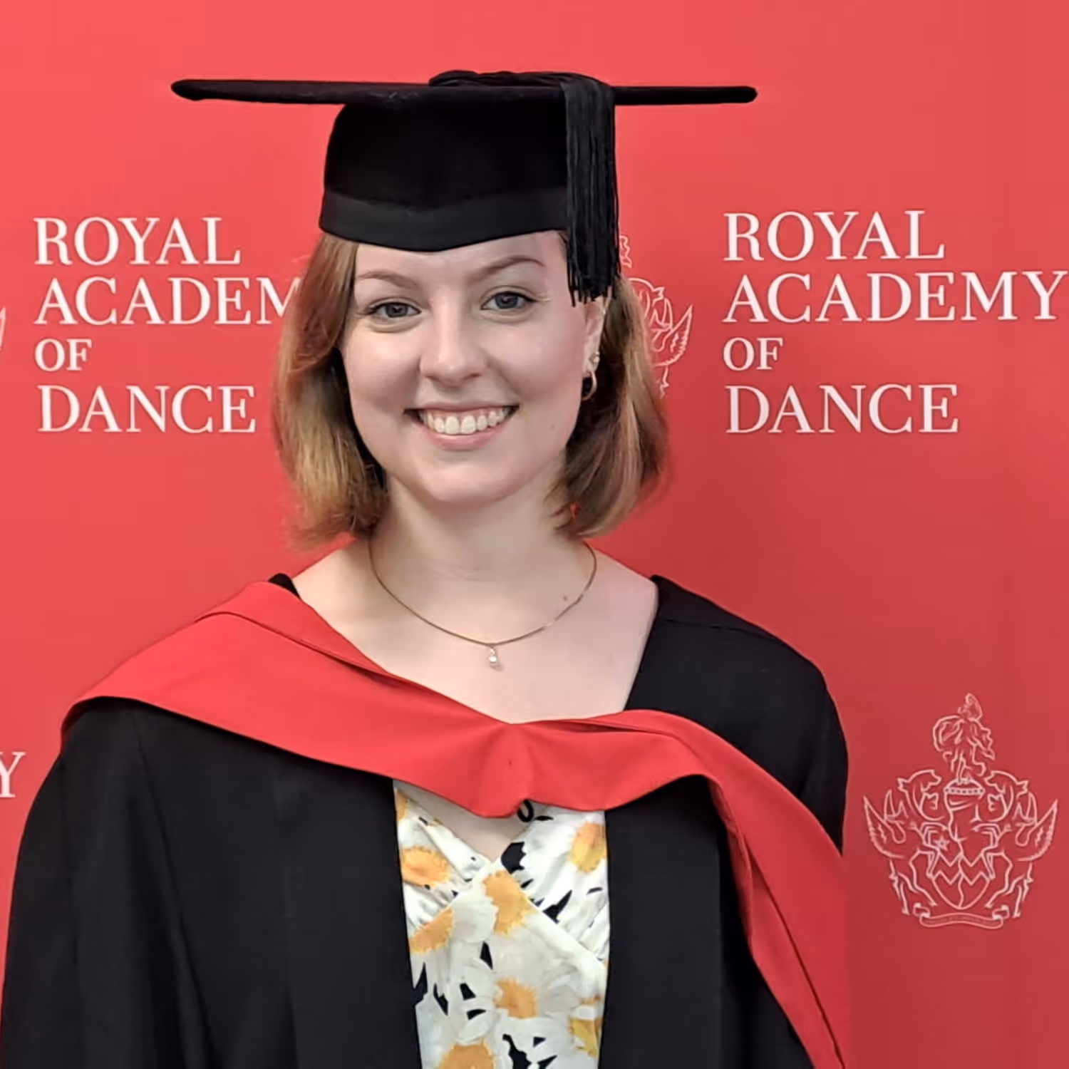 Junge Frau lächelnd in akademischem Abschlusskleid und Barett vor rotem Hintergrund mit Schriftzug Royal Academy of Dance.