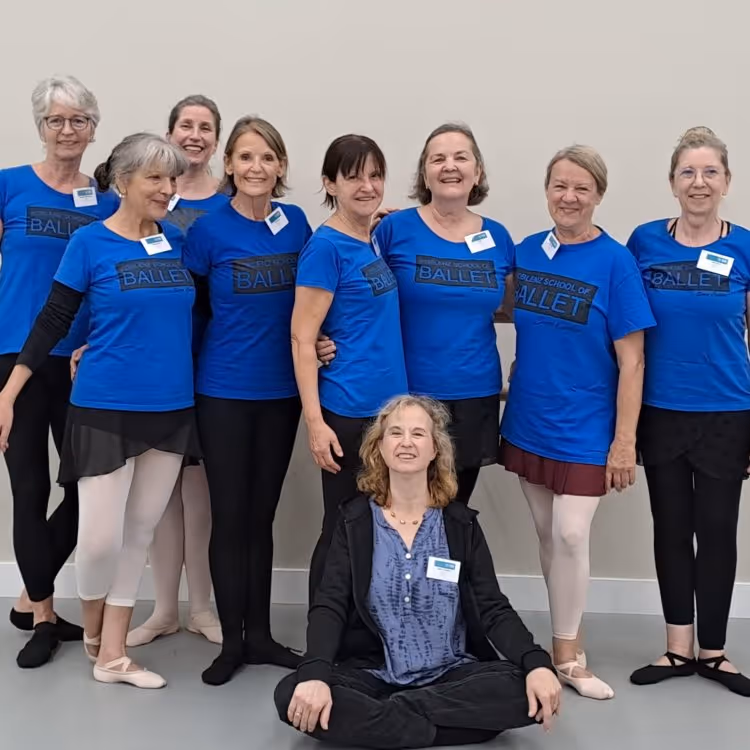 Acht Frauen in blauen T-Shirts mit dem Schriftzug 'Ballet', einige tragen Ballettschuhe und -strumpfhosen, eine Frau sitzt vorne im Schneidersitz, lächelnd.