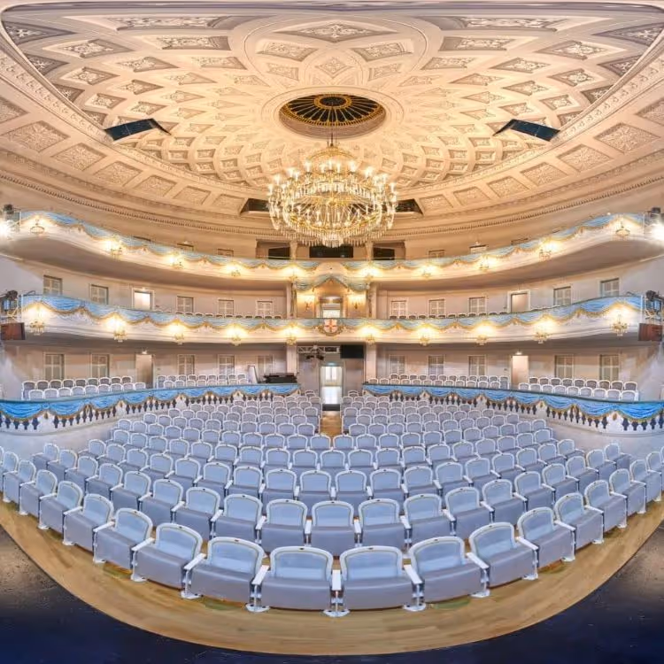 Prächtiger Theatersaal mit blauen Sitzen, verzierter Kuppeldecke und großer Kronleuchter.